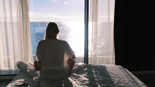 Rear View of a Woman Sitting on a Bed Opposite a Panarum Window Overlooking the Sea on a Sunny Day alt
