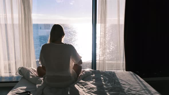 Rear View of a Woman Sitting on a Bed Opposite a Panarum Window Overlooking the Sea on a Sunny Day
