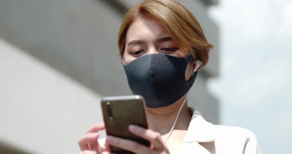Asian woman wearing earphones and face mask using mobile phone in the city