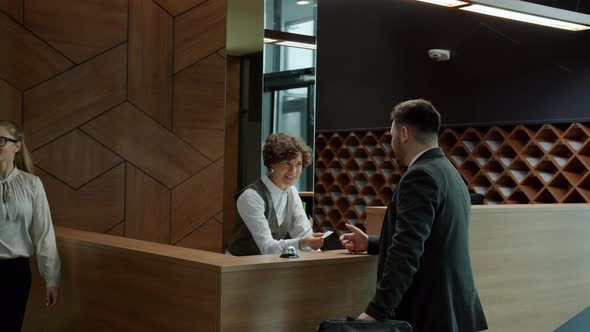 Male Guest Talking To Hotel Receptionist and Getting Key Card While Man and Woman Walking By alt