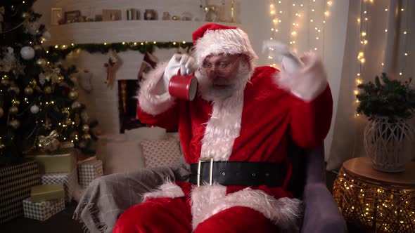 Funny Santa Claus Sits in a Chair and Jokes with a Cup Near the Christmas Tree and Fireplace
