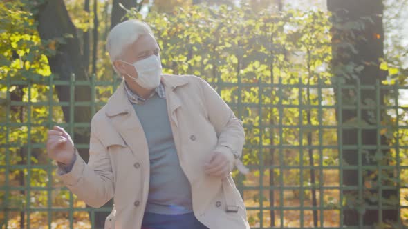 Portrait of Cheerful Grandfather in Medical Mask Dancing in Autumn Park Over Green Fence Background alt