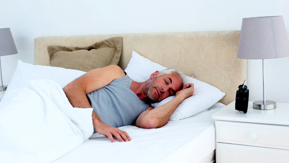 Tired Man Waking Up And Looking At His Alarm Clock alt