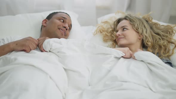 Young Happy Millennial Couple Hiding Under White Blanket Cuddling Lying in Bed alt