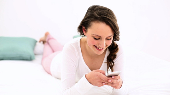 Smiling Woman Lying On Bed Texting On The Phone 1