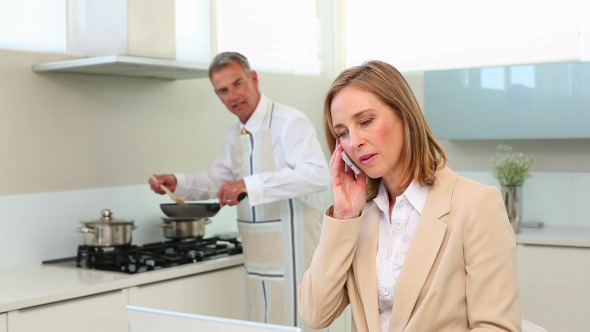 Businesswoman Talking On Phone While Husband Cooks