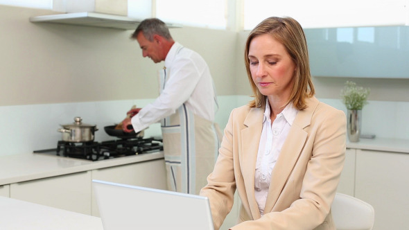 Businesswoman Using Laptop While Husband Cooks