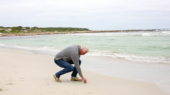 Retired Man Kneeling On The Beach Looking Out