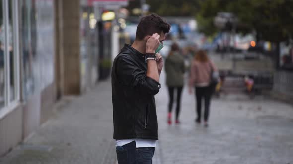 A young man in a leatherette jacket and jeans with a modern hairstyle on a busy street and puts on a alt