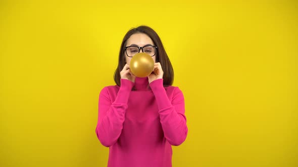 A Young Woman Inflates a Gold Balloon with Her Mouth on a Yellow Background. Girl in a Pink alt
