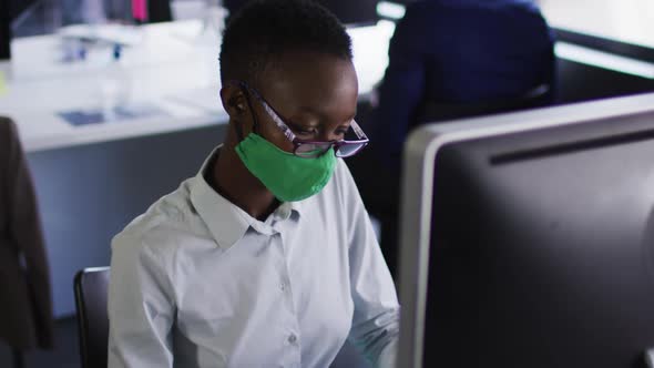 African american woman wearing face mask using computer while sitting on her desk at modern office alt