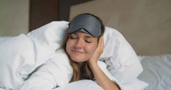 Smiling Woman in Sleep Mask Lies Under Duvet on Bed Closeup
