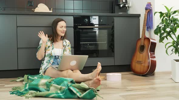 Woman in earphones listening to music using laptop sitting on floor at kitchen. Quarantine concept alt