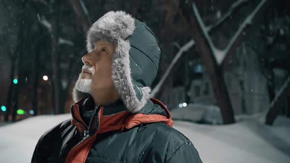 An Elderly Man with a Gray Beard in a Winter Hat is Standing on the Street at Night in the City and