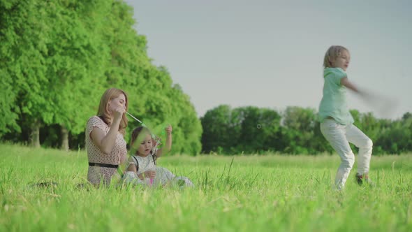 Wide Shot of Cheerful Young Mother and Cute Daughter Blowing Soap Bubbles and Blond Joyful Son alt