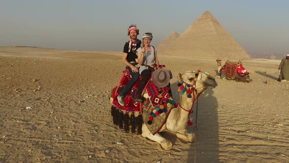 Smiling couple sitting on camel in front of Giza pyramids alt