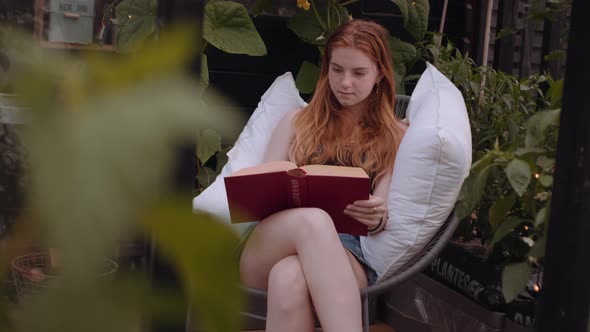 Teenage Girl Sitting On Garden Chair With Pillows And Reading alt