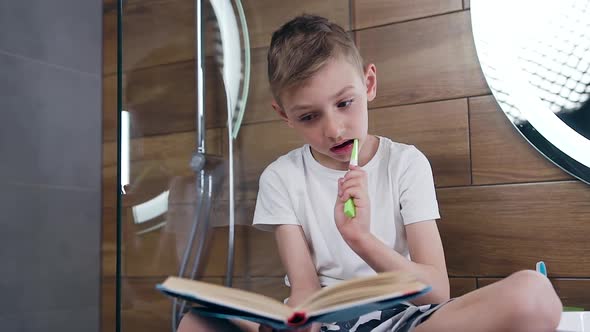 Boy Sitting Near the Bathroom Mirror Simultaneously Reading Book and Carefully Brushing Teeth alt
