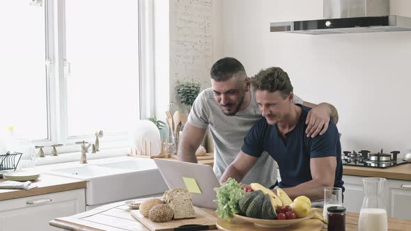 Happy gay male couple browsing internet together in kitchen at home