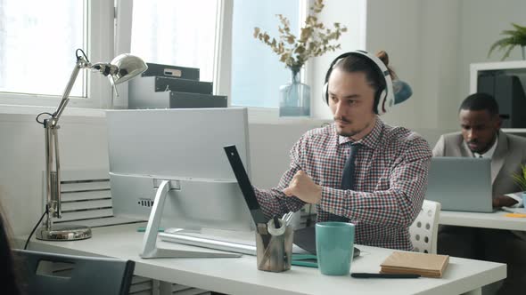 Creative Young Man in Headphones Dancing Working with Computer at Desk in Office alt