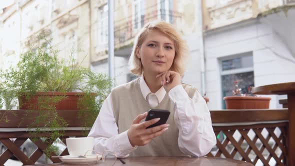 Young Blonde Woman with Elegant Manicure in a Beige Vest and White Blouse Holding a Cell Phone While alt