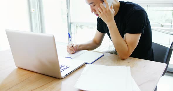 Business executive writing on diary while talking on mobile phone alt