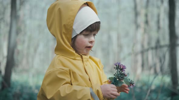 Child in Yellow Raincoat with Hood Stands in Forest and Holds Spring Flowers