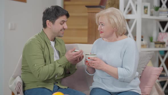 Positive Adult Son Talking with Senior Mother Holding Herbal Tea Cups alt