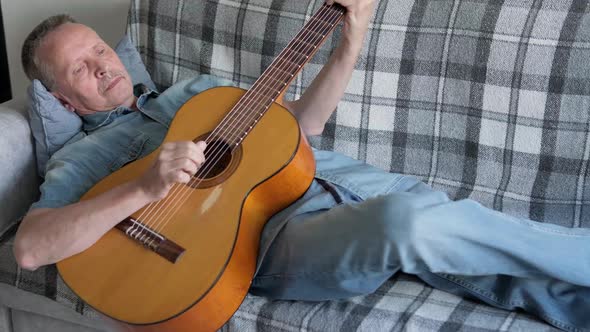 Elderly Man Playing an Acoustic Guitar at Home Lying on Couch alt
