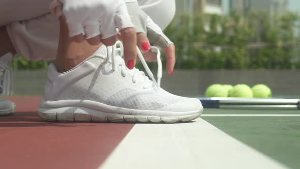 Woman's Hands Tying Shoelaces on White Shoes with Tennis Balls in Background alt