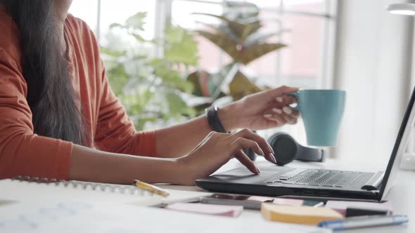 Close up hands of a businesswoman using a laptop and drinking coffee while sitting in the office.