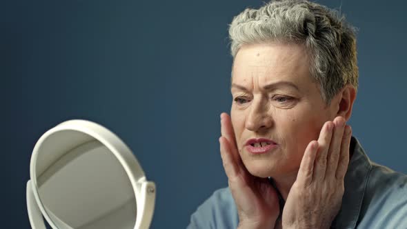 An Elderly Grayhaired Woman Carefully Examines Her Reflection in the Mirror Lightly Touching the alt