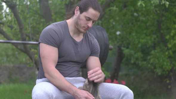 Male Athlete Sits on a Sports Simulator in His Yard and Strokes a Cat alt