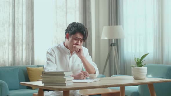 Asian Man Student Being Sleepy While Writing In The Notebook On The Table At Home alt