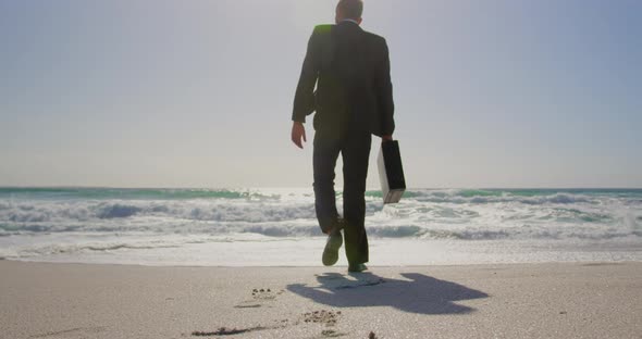 Businessman walking towards sea on beach in the sunshine 4k