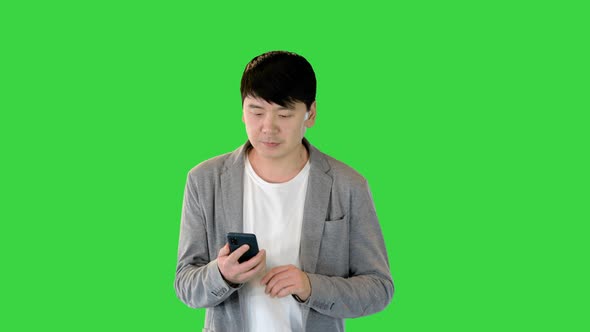 Young Asian Man Walks Looking at His Smartphone on a Green Screen Chroma Key alt