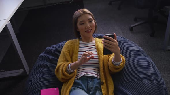 Happy asian business woman lying on bean bag using smartphone during break alt
