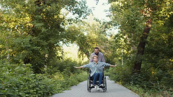 Loving couple is fooling around in park, man is rolling disabled woman in wheelchair alt
