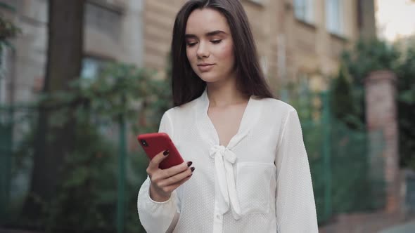 Beautiful Smiling Brunette Girl Walking Down Cosy Old Town Street While Using Her Phone and Looking alt