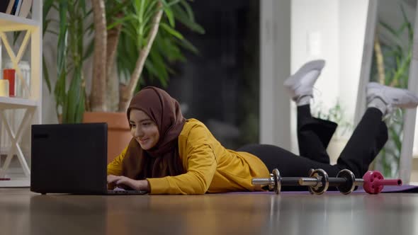 Charming Young Fit Woman in Hijab Messaging Online on Laptop After Training at Home alt