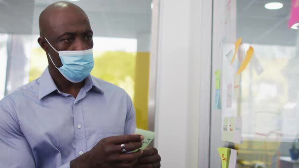 African american businessman in face mask brainstorming, putting memo notes on board in office alt