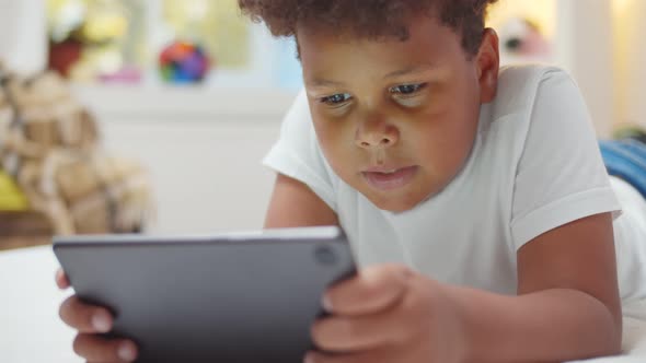 Afro-american Boy Kid Lying on Bed Watching Cartoons on Digital Tablet alt