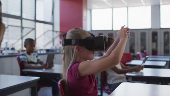 Caucasian schoolgirl sitting at desk in classroom wearing virtual reality glasses and gesticulating alt