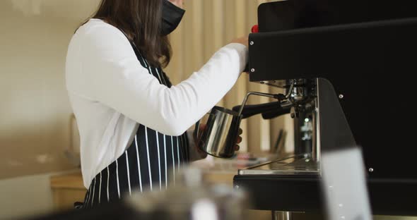 Caucasian waitress wearing face mask standing at coffee machine, preparing froth milk alt