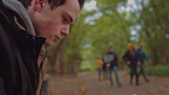 Young Guy with a Phone in His Hands Sitting on a Bench in the Park. In the alt