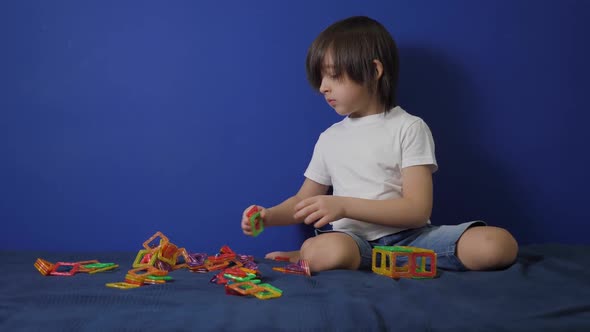 Boy Child in a White Tshirt is Sitting on a Bed Against a Blue Wall and Playing alt