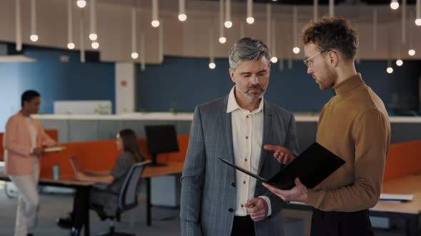 Caucasian Young Guy and Senior Man Standing Indoors at Office Center Smiling and Talking alt