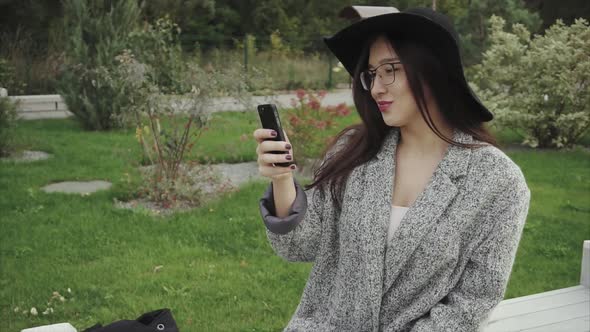 Young Attractive Woman Sits on Bench in the Park and Works on the Smartphone alt