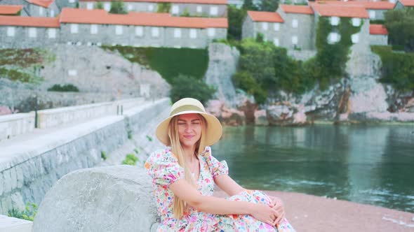 Zoom Shot of Charming Blonde Woman Sitting on the Bank in Hat and Dress