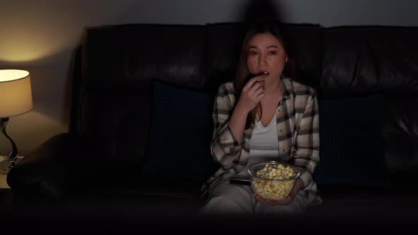 young woman watching TV and eating popcorn on sofa at night